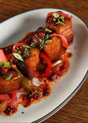 A close-up shot of a plated dish featuring glazed pork belly cubes topped with red chili powder, scallions, and green herbs, served on a white oval plate with a dark rim on a wooden table.