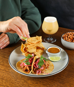 Person adding a lime wedge to tacos on a plate with a glass of beer and bowl of beans in the background.