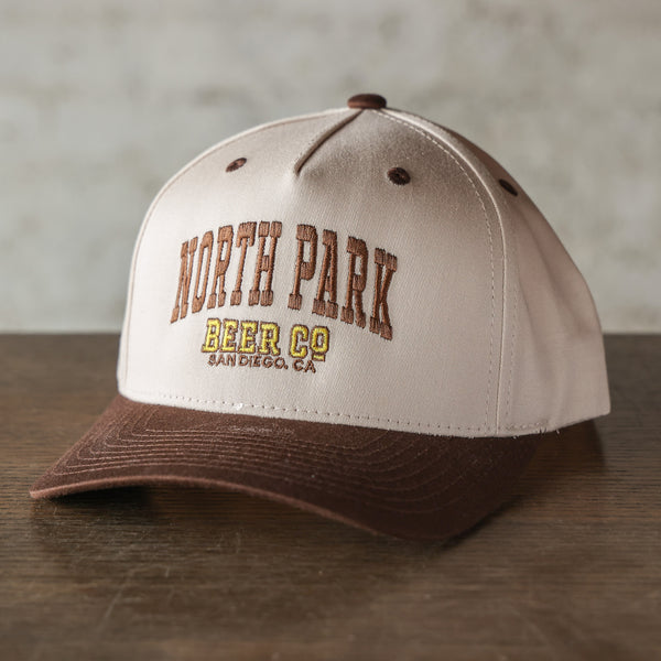 A structured baseball cap with a cream-colored crown and a brown brim, featuring 'NORTH PARK BEER CO SAN DIEGO, CA' embroidered on the front. The cap is displayed on a dark wooden surface against a neutral, textured gray background.