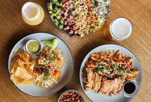 Overhead view of a restaurant table with two plated meals and two beer glasses on a wooden table. One plate has tacos with tortilla chips, guacamole, lime, and salsa, and the other has pulled pork with cheese fries, cilantro, and salsa.