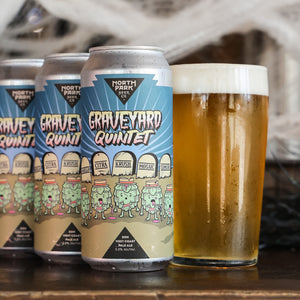 Three aluminum cans of North Park Brewing Co. Graveyard Quintet beer lined up on a wooden surface next to a glass filled with golden beer and white foam, with a blurred rustic background.