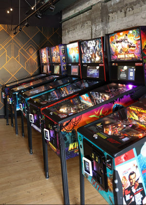 A row of colorful pinball machines lined up in an arcade or entertainment venue with a concrete wall, wood floor, and geometric patterned accent wall.