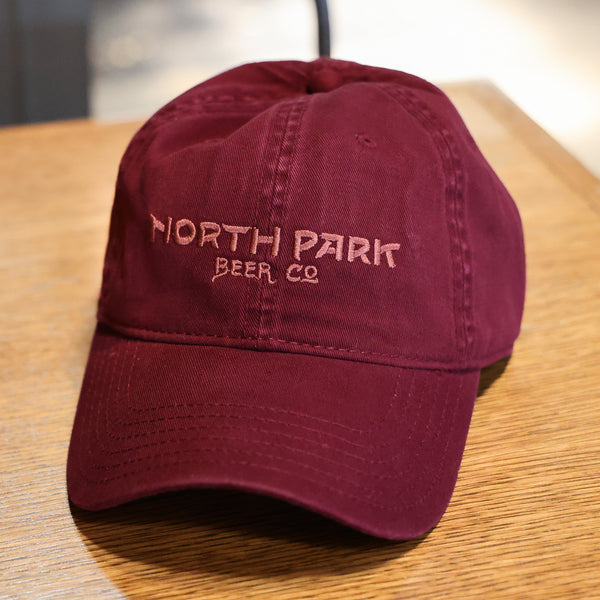 Maroon Dad Hat with North Park Beer Co logo embroidered on the front in a tonal maroon color, sitting on a table.