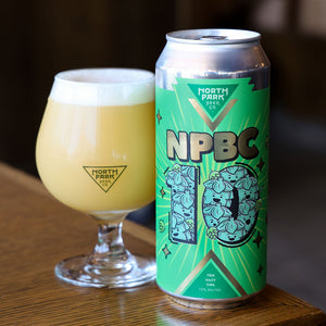 A can of North Park Beer Co. NPBC 10 Year Hazy TIPA next to a filled tulip glass with a foamy head, set on a wooden table.