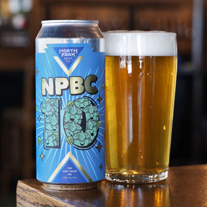A can of North Park Beer Co NPBC 10 West Coast IPA next to a glass of golden beer with white foam, set on a wooden bar table.