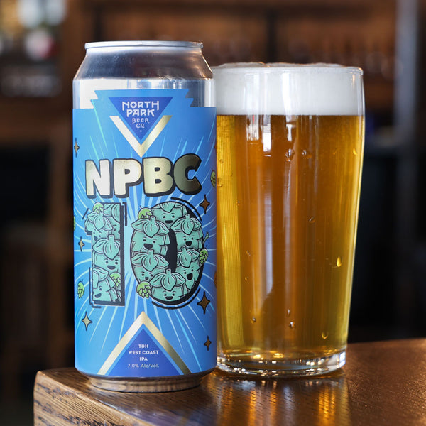 A can of North Park Beer Co NPBC 10 West Coast IPA next to a glass of golden beer with white foam, set on a wooden bar table.