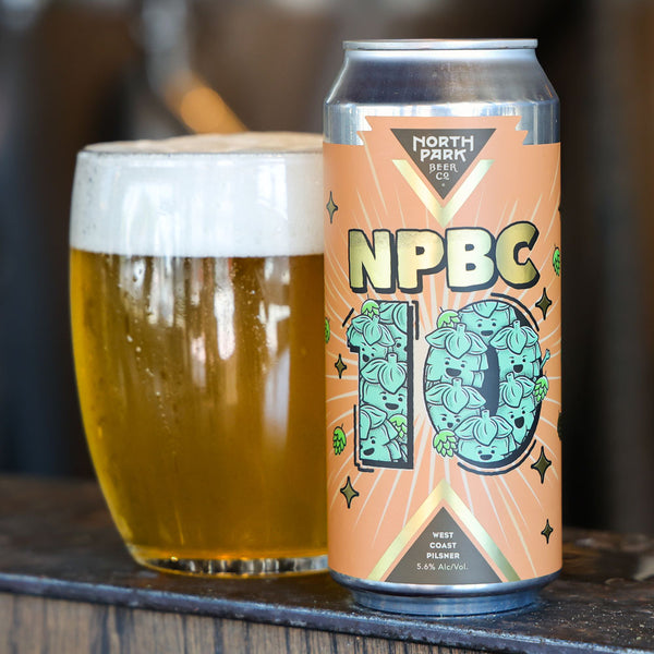 A can of North Park Beer NPBC 10 West Coast Pilsner next to a glass of golden beer with white foam, set on a dark wooden surface.