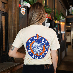 Someone wearing a tshirt featuring our Simcoe Slammer mascot on the back in orange, navy and light blue on a beige tshirt.