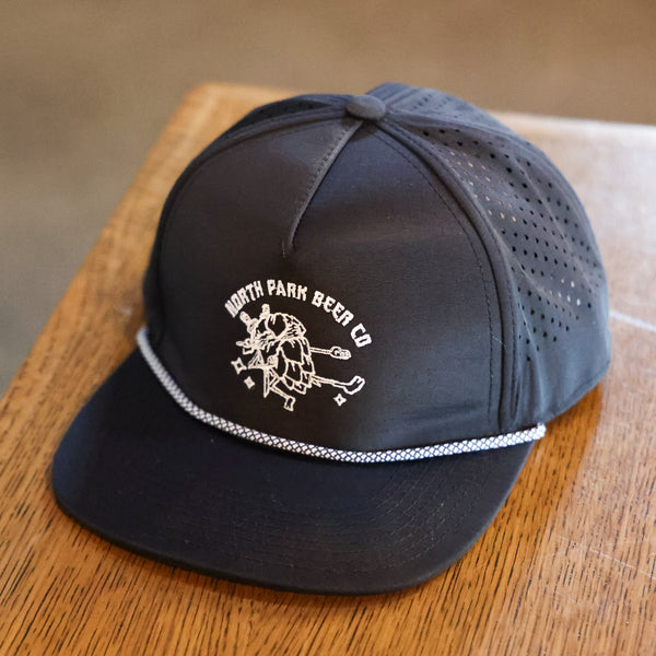 Black Laser Cut Nylon hat with our Hop-Fu! logo embroidered in white on the front. Also a white rope along the top of the bill, is sitting on a tabletop.