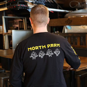 Someone wearing a Black Long Sleeve Tshirt featuring our Art is Hard Buddies across the back and North Park logo above it in yellow and white.