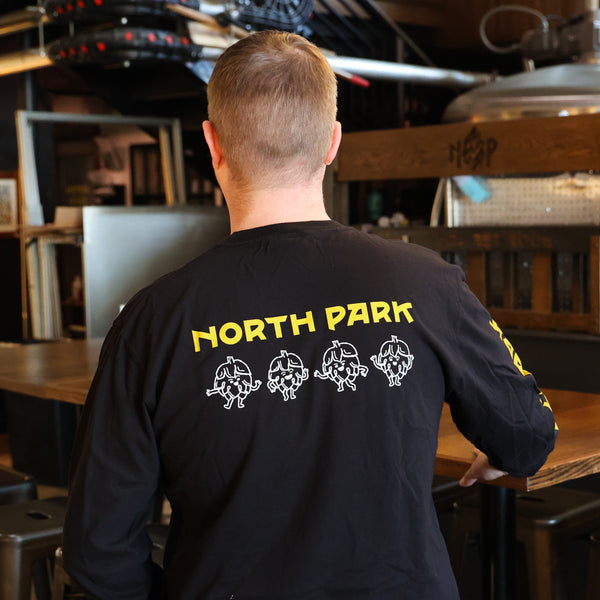 Someone wearing a Black Long Sleeve Tshirt featuring our Art is Hard Buddies across the back and North Park logo above it in yellow and white.