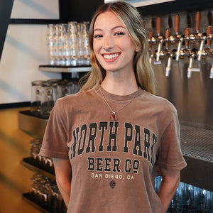 Someone wearing a tshirt featuring NPBC Collegiate Style Logo in brown and beige on the front of an espresso brown tshirt.