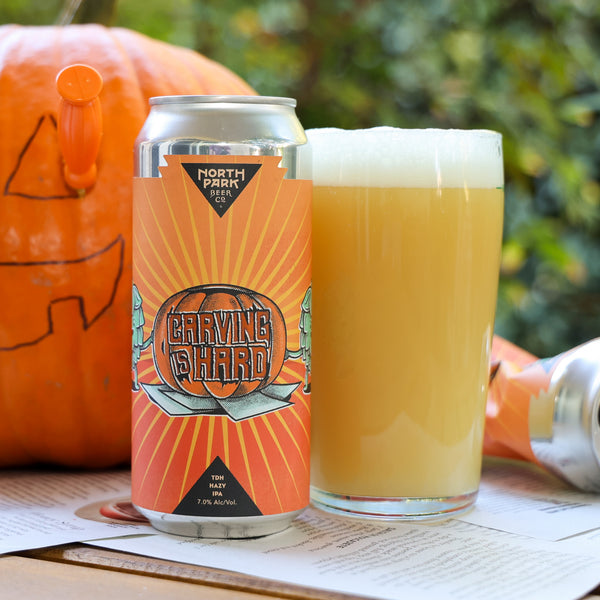 A can of North Park Brewing Co. Carving Hard beer stands next to a filled pint glass with foam, set outdoors on a table with an orange pumpkin and blurred green foliage in the background.