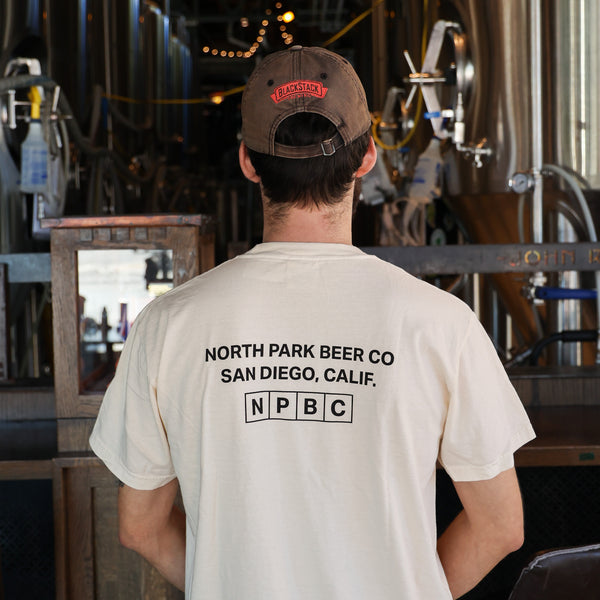 A man viewed from behind wearing a cream-colored t-shirt with 'NORTH PARK BEER CO SAN DIEGO, CALIF. NPBC' printed in black on the back in a brewery setting.