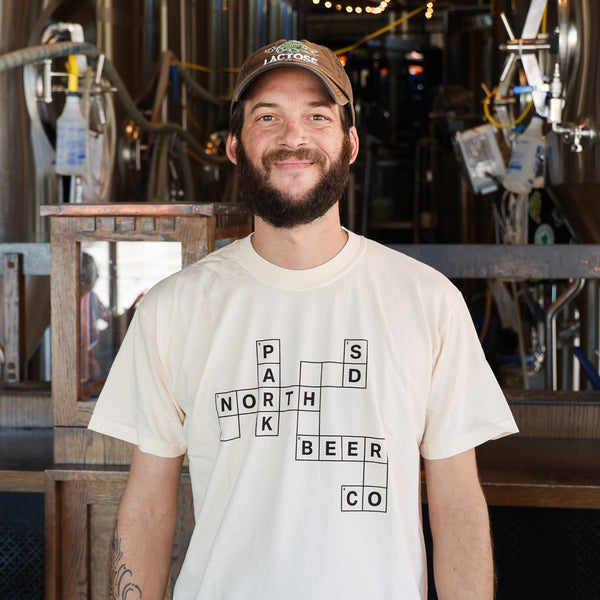 A smiling man stands in a brewery wearing a cream-colored t-shirt featuring a crossword puzzle graphic spelling out ' NORTH PARK BEER CO and SD'.