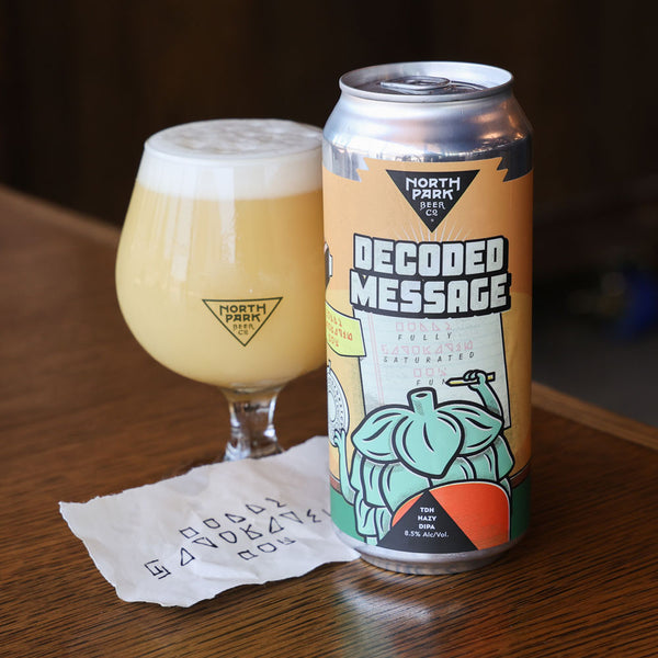 A can of North Park Beer 'Decoded Message' next to a tulip glass of pale golden beer with foam, on a wooden table with a handwritten paper note.