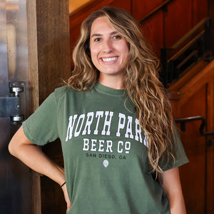 Someone wearing a green 'North Park Beer Co' Collegiate Logo t-shirt in a brewery setting