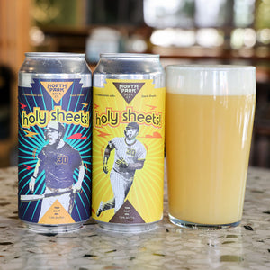 Two North Park Beer Co beer cans labeled 'Holy Sheets!' stand on a countertop beside a filled pint glass of pale yellow beer, with a blurred indoor background.