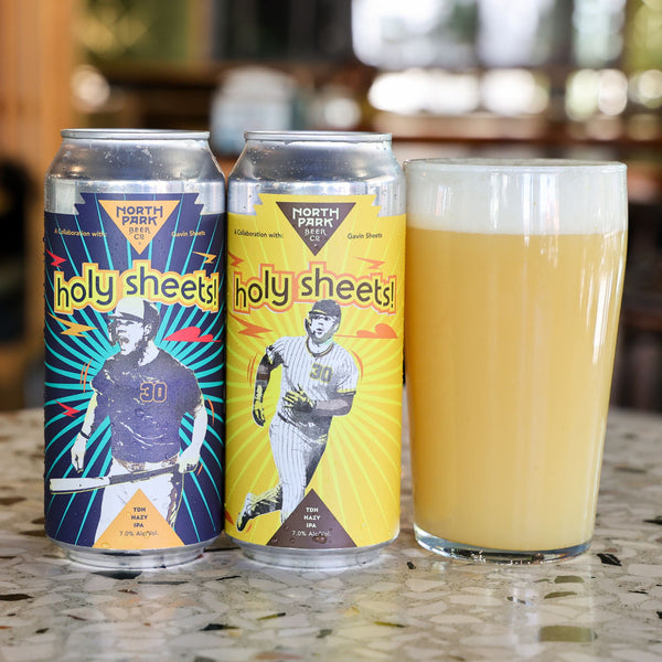 Two North Park Beer Co beer cans labeled 'Holy Sheets!' stand on a countertop beside a filled pint glass of pale yellow beer, with a blurred indoor background.