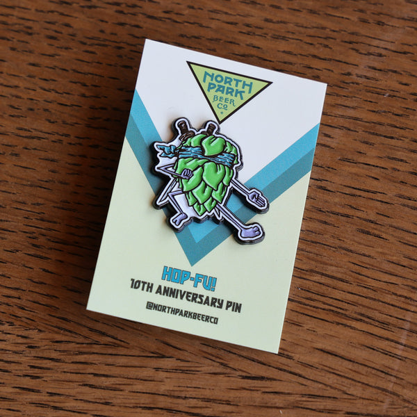 A Hop-Fu! North Park Beer Co. enamel pin on its original backing card, placed on a wooden table.