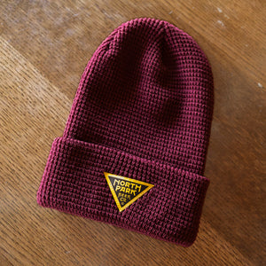 Purple waffle knitbeanie with gold and black triangle logo patch.  Shown flat on a table.