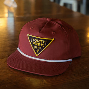 Maroon Cotton/Poly 5 panel hat with a triangle logo patch on the front and a rope detail across the bill. Sitting on a table.