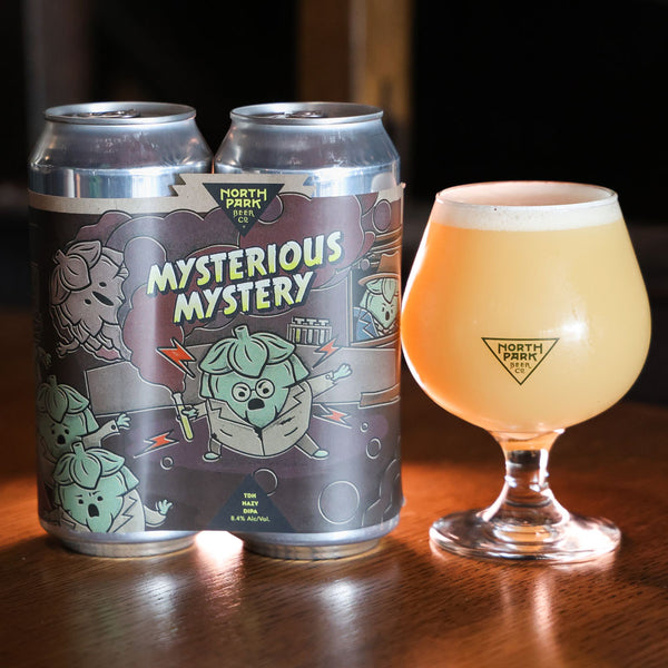 Two cans of 'Mysterious Mystery' beer from North Park Beer Co. next to a glass of beer on a wooden surface.