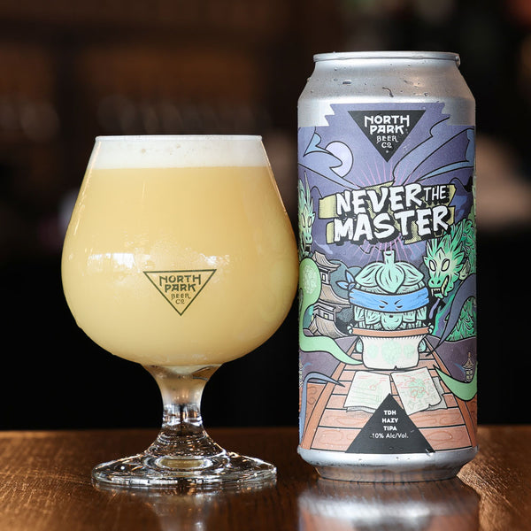 A glass of pale yellow beer with white foam sits beside a silver can of North Park Beer labeled 'Never The Master' on a wooden table against a dark blurred background.