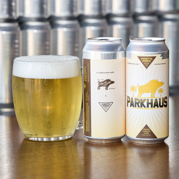 Two cans of Parkhaus beer and a glass of beer on a wooden surface with metal kegs in the background.