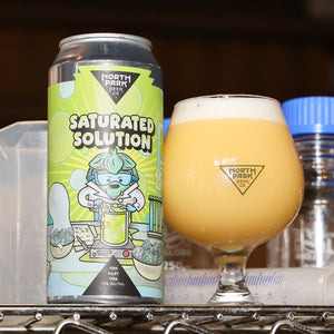 A can of North Park Brewing Saturated Solution IPA next to a tulip glass filled with the golden beer and white foam, set on a metal shelf.