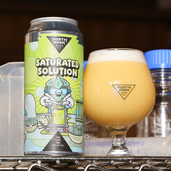 A can of North Park Brewing Saturated Solution IPA next to a tulip glass filled with the golden beer and white foam, set on a metal shelf.