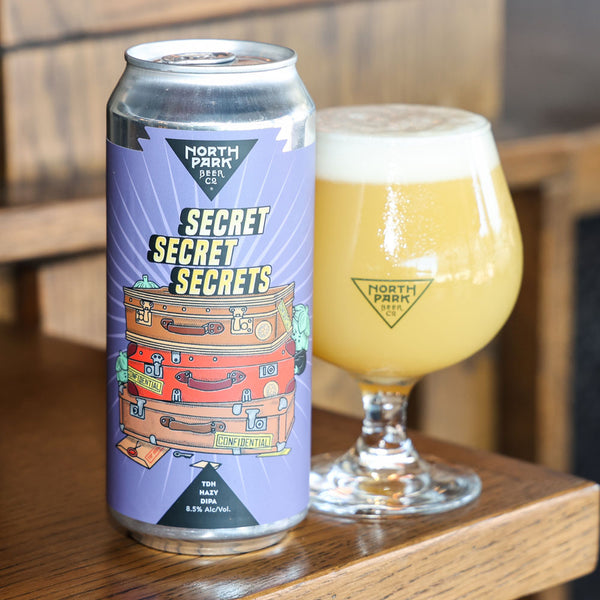 A can of North Park Beer 'Secret Secret Secrets' stands on a wooden table next to a filled tulip glass of the beer, with a softly blurred wooden background.