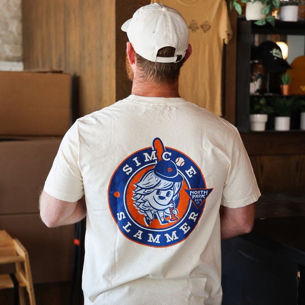 Someone wearing  a beige T-shirt with our Simcoe Slammer logo on the back in navy, light blue and orange.