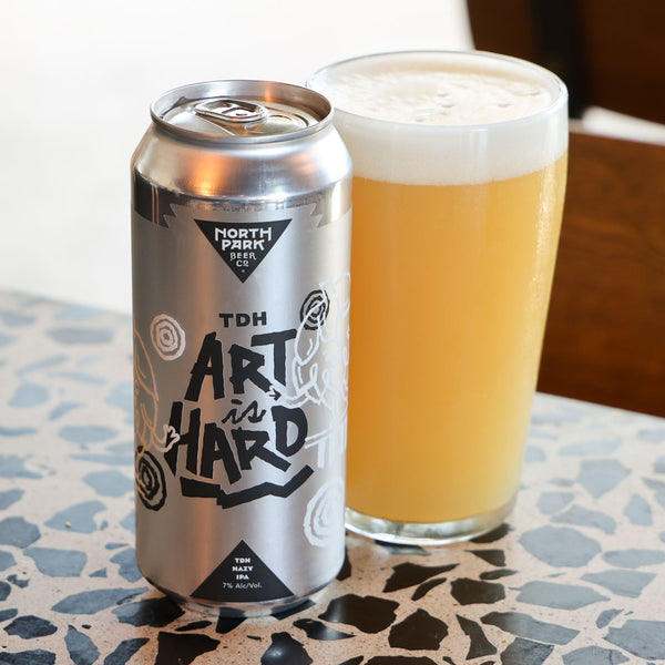 A silver aluminum can of North Park Beer Co TDH Art Is Hard beer stands next to a pint glass filled with golden beer and white foam, set on a speckled stone surface.