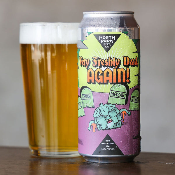 A can of North Park West Coast 'Very Freshly Dead Again!' IPA stands on a wooden table next to a glass of golden beer with white foam, against a neutral gray background.