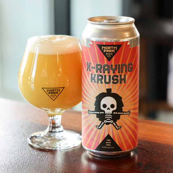 Can of North Park Beer X-Raying Krush next to a glass of beer on a wooden surface.