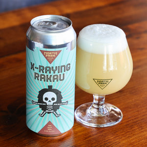 A can of North Park Beer Co. X-Raying Rakau next to a filled tulip glass of the pale golden beer on a wooden table.